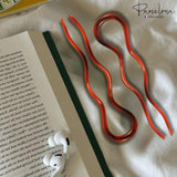 Two brown hairpins on an open book with 'Parcelona' branding in the corner.