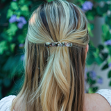 Person with long blonde hair tied back with a gray magma hair clip, blurred green and purple flowers in the background